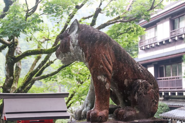 a stone cat covered in moss facing away towards a tree and a building behind it, thumbnail
